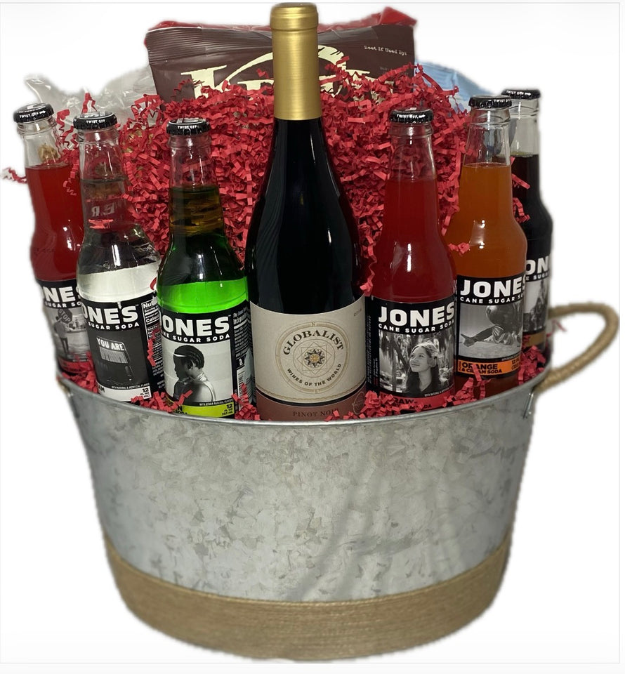A metal gift basket filled with assorted bottled drinks and a wine bottle, arranged in red shredded paper. The basket has a galvanized metal body, a natural jute rope handle, and a rope-wrapped base.