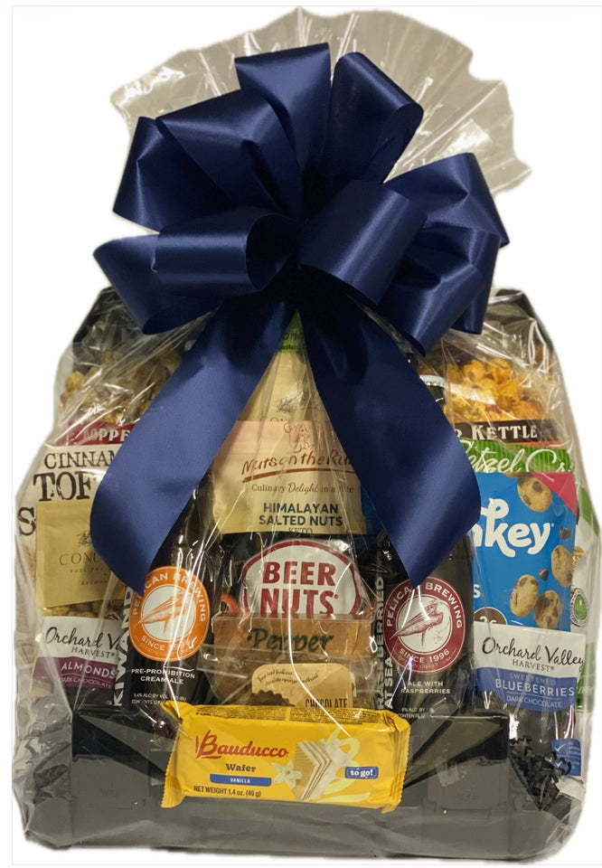 A gourmet gift basket filled with assorted snacks, nuts, and packaged foods, wrapped in clear cellophane and tied with a large navy blue satin bow. Visible items include Bauducco chocolate, Mountain Dew, Himalayan salted nuts, and Orchard Villa crackers.