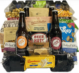 A curated snack and beer gift basket arranged in a black plastic tray, featuring two bottles of Oregon Brown Ale, several bags of nuts and chocolates, and packaged treats from brands like Conundrum and Baudoucci.